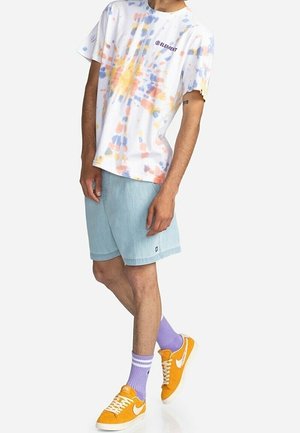 Osoba nosząca białą koszulkę Element z efektem farbowania nawiązującym do tie-dye, jasnoniebieskie krótkie spodenki, fioletowe skarpetki oraz pomarańczowe buty Nike, stojąca z jedną nogą skrzyżowaną.