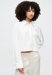 White cropped button-up shirt with long sleeves and a pointed collar, made of smooth fabric. Paired with light beige pants.