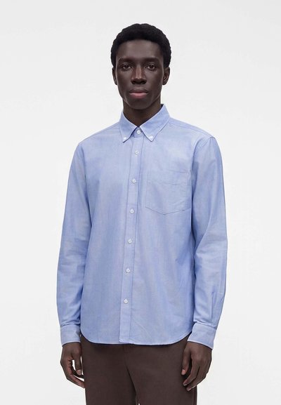 Homme portant une chemise bleu clair à boutons avec une poche poitrine et un pantalon marron, debout devant un fond blanc uni.