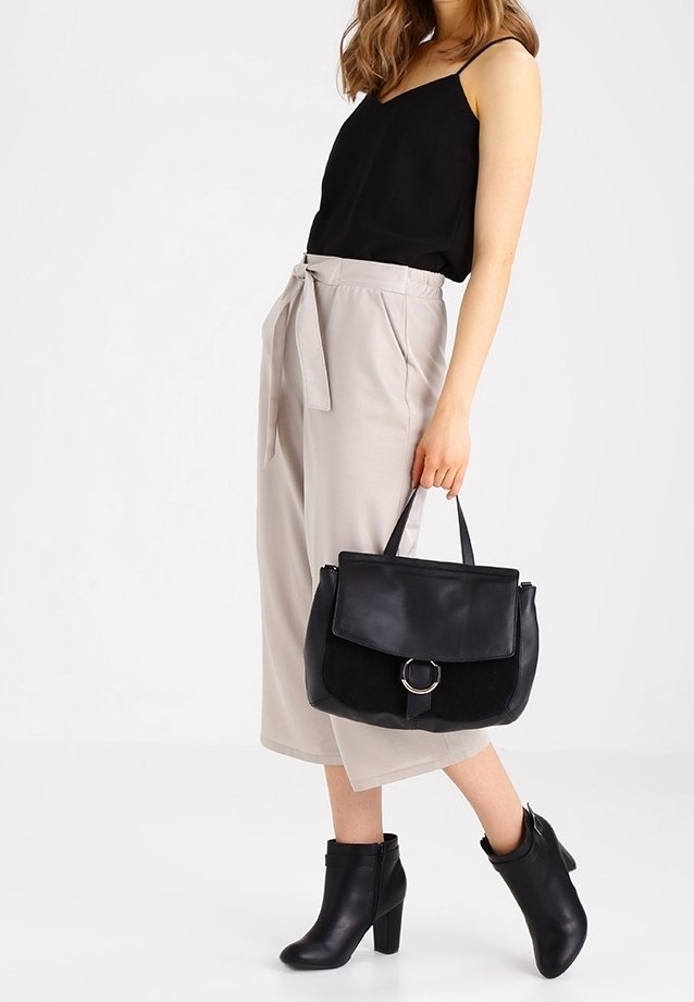 Femme portant un haut noir sans manches, pantalon beige large et court, bottines noires à talons, tenant un sac à main noir avec un détail d'anneau argenté.