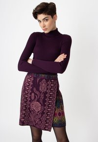 Rippstrukturierter dunkelvioletter Rollkragenpullover kombiniert mit einem knielangen Rock, der florale und geometrische Muster in Lila, Pink und Grün aufweist.