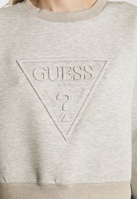 Sweat-shirt gris clair avec logo triangulaire Guess embossé, comportant "GUESS", "U.S.A" et un grand point d'interrogation au centre.