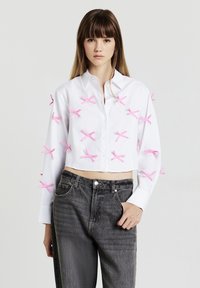 Weiße, kurz geschnittene Bluse mit mehreren rosa Schleifenakzenten an den Ärmeln, kombiniert mit dunkelgrauen, hoch taillierten Jeans.