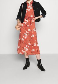 Abito midi floreale in rame con bottoni e vita regolabile, abbinato a una giacca di jeans nera e stivaletti neri. Inclusa una piccola borsa rosa.