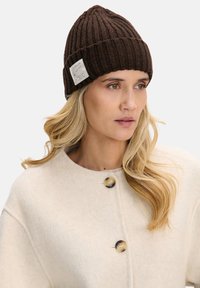 Brązowa prążkowana dzianinowa czapka z podwiniętym mankietem i tkana etykietą na boku. Miękka tekstura, dopasowany kształt i ciepły design.