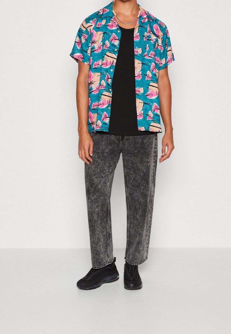 Personne portant une chemise à manches courtes à fleurs couleur sarcelle sur un t-shirt noir, un jean gris délavé, des baskets noires et un collier chaîne en argent.