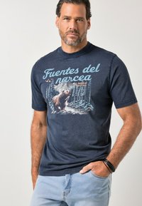 Man in een marineblauw T-shirt met de tekst "Fuentes del Narcea" en een afbeelding van een beer in een bos, gecombineerd met lichtblauwe spijkerbroek en zwarte armbanden.
