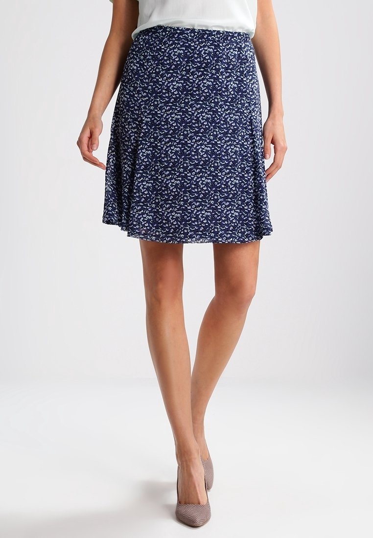 Woman wearing a navy blue floral A-line skirt and beige high heels, standing against a plain white background.