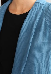 Detailní záběr horní části trupu oblečeného v černém topu a jemně modré rebrované kardiganu s šálovým límcem.