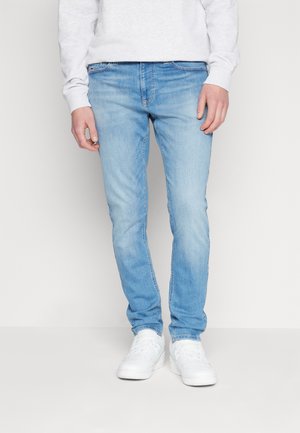 Vaqueros de mezclilla azul claro con corte slim, que presentan una textura suave, un ligero desvanecimiento y un diseño tradicional de cinco bolsillos, combinados con zapatillas blancas.
