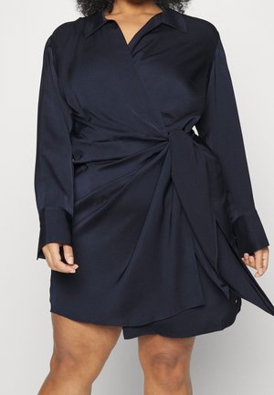 Czarna sukienka kopertowa z długim rękawem, kołnierzykiem i wiązaniem z boku, noszona przez osobę o średnim odcieniu skóry, widoczna od szyi do połowy uda.