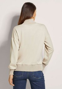 Lichtbeige trui met een hoge hals, geribbelde mouwen en zoom; zachte textuur en een relaxed fit, gecombineerd met donkerblauwe jeans.