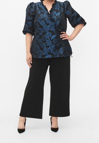 Donna che indossa una blusa a fiori blu con maniche a sbuffo, pantaloni larghi neri e tacchi a punta neri, in piedi con una mano sul fianco.