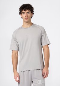 Młody mężczyzna z kręconymi włosami, ubrany w jasnoszary sportowy t-shirt z krótkim rękawem i pasujące szorty, na jednolitym białym tle.