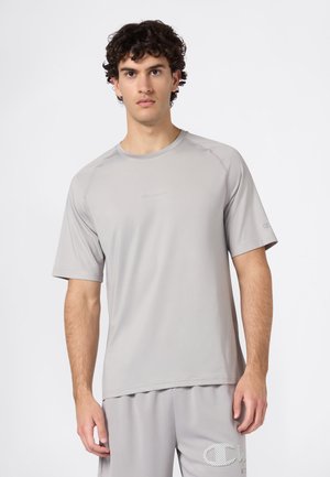Jonge man met krullend haar die een lichtgrijs sportshirt met korte mouwen en bijpassende shorts draagt, tegen een plain witte achtergrond.