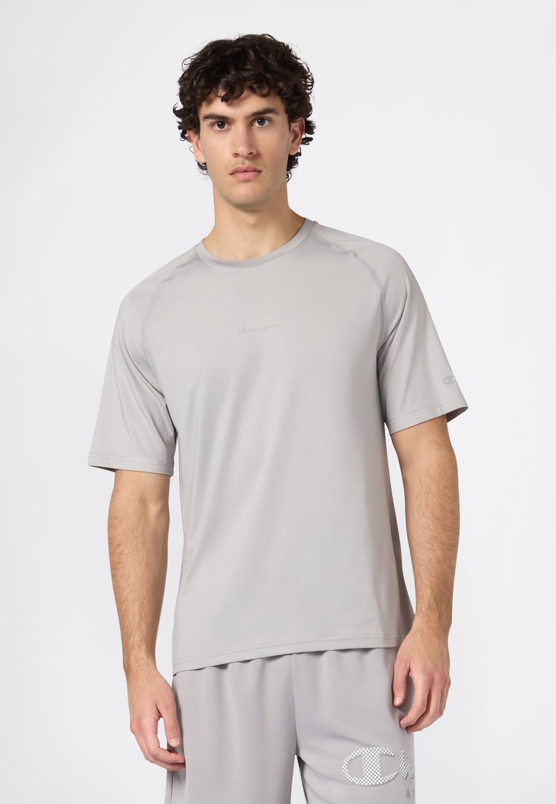 Młody mężczyzna z kręconymi włosami, ubrany w jasnoszary sportowy t-shirt z krótkim rękawem i pasujące szorty, na jednolitym białym tle.