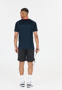 Marinblå idrottströja med röd krage, kombinerad med svarta shorts. Modellen håller en racket, bär vita strumpor och svarta och vita skor.