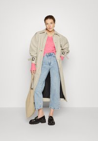 Langer karierteter Trenchcoat mit übergroßen Ärmeln über einem leuchtend pinken Oberteil, kombiniert mit hellblauen weiten Jeans und schwarzen klobigen Mokassins.