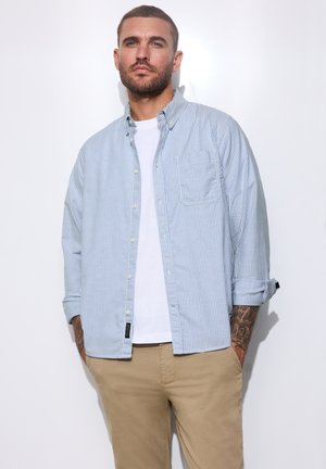 Mann mit kurzen Haaren und Bart, der ein blau gestreiftes Hemd über einem weißen T-Shirt und beige Hose trägt, steht mit den Händen in den Taschen.