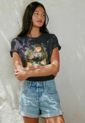 Junge Frau mit langen dunklen Haaren trägt ein schwarzes T-Shirt mit tropischem Grafikaufdruck und hochgekrempelte helle Jeansshorts vor einer strukturierten weißen Wand.