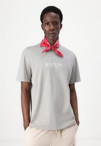 Camiseta de algodón gris con el logo "REPLAY" en blanco y el año "1981". Se lleva con una bufanda de bandana roja, añadiendo un toque de color.