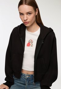 Felpa nera con cappuccio e tasche frontali, abbinata a una t-shirt bianca corta con un design ricamato di un cane sul petto.