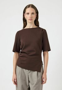 Top corto marrone a maniche corte in tessuto liscio, con scollatura discreta, fessure laterali e silhouette aderente, abbinato a pantaloni grigi chiari.