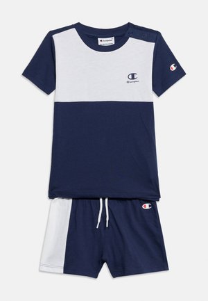 Småbørns navyblå og hvide Champion-sæt med korte ærmer, med logo på bryst, ærme og shorts, med trykknapper på skulderen og snøre i taljen.