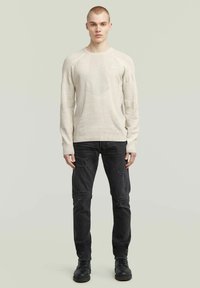 Beiger Strickpullover mit strukturiertem Muster, Rundhalsausschnitt und gerippten Bündchen, kombiniert mit schwarzen, ausgewaschenen Jeans und schwarzen Schnürstiefeln.