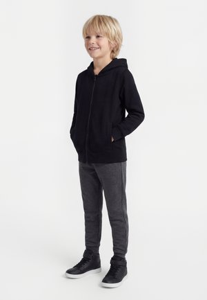 Usmievajúce sa blond dieťa stojace s rukami v vreckách, oblečené v čiernej mikine na zips, sivých teplákoch a čiernych vysokých teniskách.
