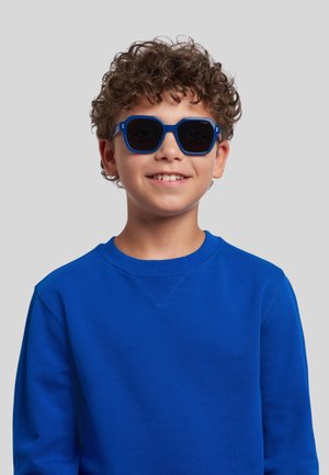 Ragazzo sorridente con capelli ricci che indossa occhiali da sole quadrati blu scuro e una felpa azzurra brillante su uno sfondo grigio uniforme.