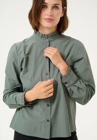 Green blouse with a high, ruffled collar, front button closure, and gathered sleeves. Made from smooth fabric with slight sheen.