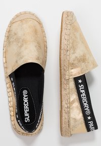 Espadrilles metálicas doradas con diseño de fácil puesta, detalle de logo elástico negro, borde de yute trenzado y un interior de tela negra.