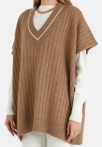 Brązowy, oversize'owy ponczo z prążkowaną fakturą, dekoltem w serek i białymi akcentami. Noszony na białej, długorękawowej koszuli z krótkimi rękawami.