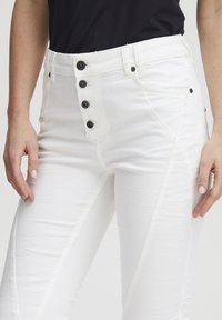 Le jean blanc présente une taille haute avec cinq boutons noirs, des poches latérales et un tissu texturé avec des détails de couture contrastants.