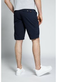 Bermuda cargo blu navy con vestibilità rilassata, chiusura con bottone e tasche laterali, abbinate a sneaker bianche su uno sfondo chiaro.