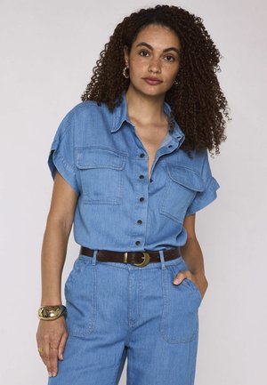 Donna con capelli ricci che indossa una camicia di jeans blu a maniche corte con bottoni e pantaloni a vita alta abbinati con cintura marrone.