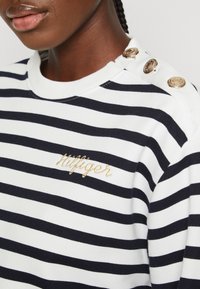 Felpa a righe bianca e blu navy, con logo ricamato in oro, collo a coste e bottoni decorativi dorati sulla spalla.