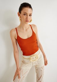 Top arancione a costine con spalline sottili, abbinato a pantaloni in crochettè crema con una cintura intrecciata dotata di un accento circolare.