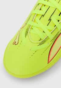 Neon gul fodboldsko med tekstureret overflade, med sorte mærker og en orange detalje. Snørebåndene er lysegule og stramt bundet.