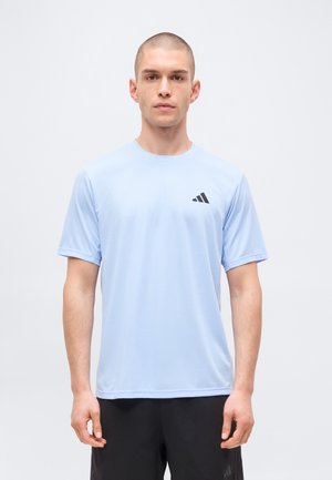 Mann mit rasiertem Kopf, der ein hellblaues kurzärmeliges Sportshirt mit schwarzem Logo und schwarzen Shorts trägt, steht vor einem einfarbigen weißen Hintergrund.