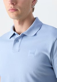 Svetlomodrá polo košeľa vyrobená z textúrovaného materiálu. Obsahuje golier, dva gombíky a nenápadné reliéfne logo na hrudi.