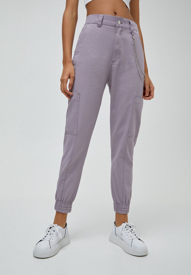 Pantalons cargo violets avec des poignets élastiques, des poches avant et un détail en chaîne argentée. Associés à des sneakers blancs sur un fond neutre.