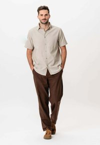 Mand der går fremad med hænderne i lommerne, iført en beige kortærmet skjorte med knapper, brune bukser og brune snørebåndssko.