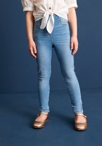 Hellblaue Skinny-Jeans mit dezenten Ausbleichungen, kombiniert mit metallic-bronzenen Ballerinas. Das Outfit wird von einem weißen Shirt ergänzt, das um die Taille gebunden ist.