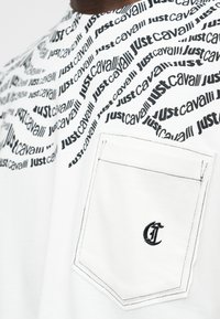 Κοντινό πλάνο λευκού πουκάμισου με μαύρα μοτίβα κειμένου "Just Cavalli" και μια μπροστινή τσέπη με μικρό μαύρο κεντητό λογότυπο.