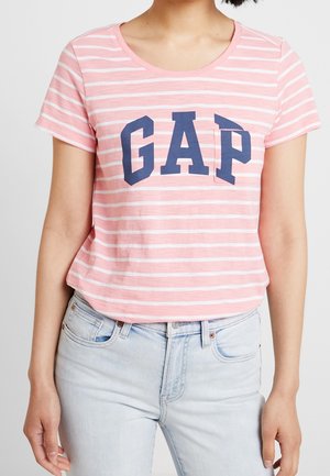Personne portant une chemise à manches courtes rayée rose et blanche avec un logo "GAP" bleu marine et un jean bleu clair, debout devant un fond uni.