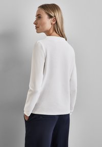 Top blanco de manga larga con un acabado texturizado y cuello redondo, combinado con pantalones azul marino. La vista trasera muestra un diseño simple y mangas ajustadas.