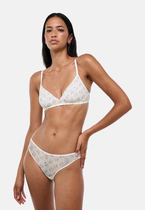 Femme aux longs cheveux foncés portant un soutien-gorge blanc à imprimé floral et une lingerie assortie, debout avec une main sur la hanche sur un fond uni.