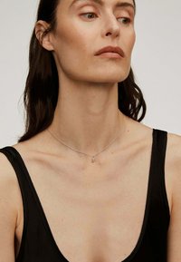 Collar de plata con un fino eslabón, que presenta un pequeño charm con un diseño geométrico; el modelo lleva una blusa negra sin mangas.
